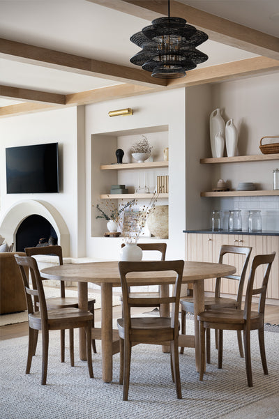 Dining room with a round wood table and chairs, arched fireplace, built-in shelves, exposed wood ceiling beams, and a black sculptural pendant light overhead.