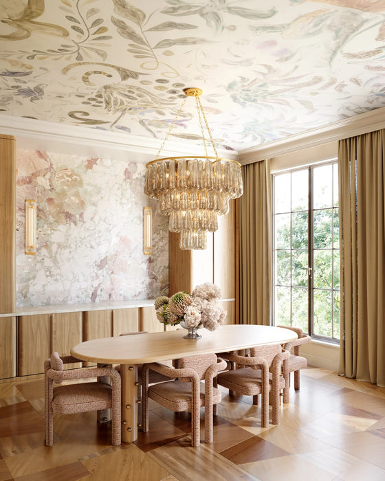 Elegant dining room featuring a tiered glass chandelier suspended from a hand-painted celestial ceiling, paired with modern sconce lighting on a marble wall, warm wood cabinetry, and a large oval dining table surrounded by upholstered chairs, with natural light streaming through tall windows.