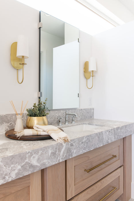 Modern bathroom vanity with a marble countertop, wood cabinetry, brass wall sconces flanking a mirror, and neutral accessories styled on the counter.