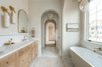 Elegant bathroom with a wood double vanity, marble countertop, brass wall sconces and fixtures, arched doorways, and a freestanding soaking tub beside a window.