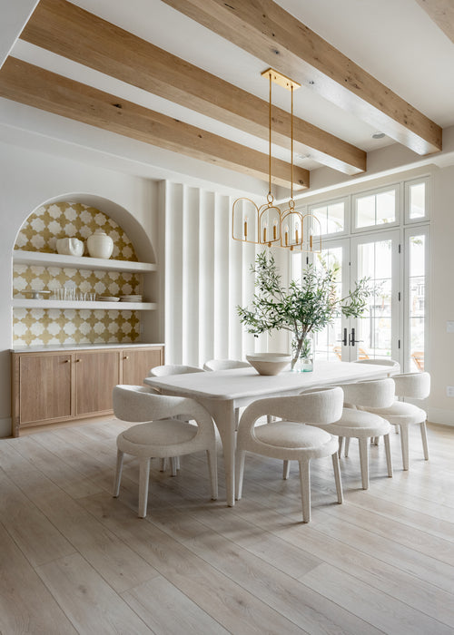 Light-filled dining room with a brass chandelier hanging above a white dining table, upholstered chairs, exposed wood ceiling beams, and an arched built-in cabinet.