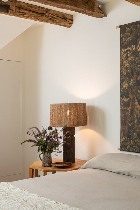 Rustic bedroom with exposed wood ceiling beams, a textured table lamp on a wood nightstand, soft neutral bedding, and floral wall art.
