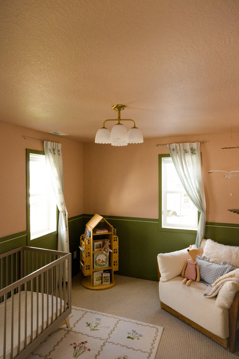 A nursery room with peach-colored walls, a green wainscoting panel, a white crib, a cream sofa with pillows, a floral rug, and a small yellow toy house organizer near the windows.