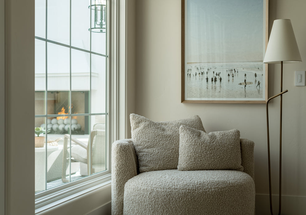 Cozy reading nook with a textured upholstered armchair beside a large window, a slim brass floor lamp, and coastal artwork on a neutral wall.