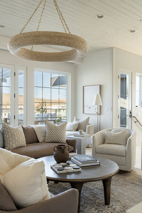 Bright living room with a woven ring chandelier suspended from a white plank ceiling above neutral sofas, armchairs, and a round wood coffee table near large windows.
