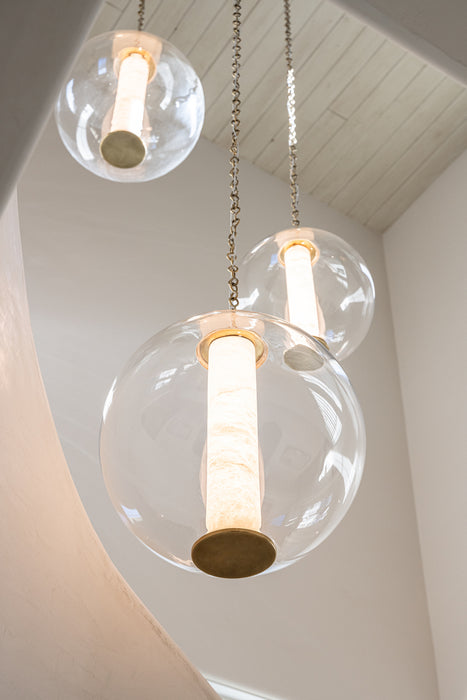 Close-up of three clear glass globe pendant lights with brass accents and glowing cylindrical light sources hanging from chains in a bright stairwell.