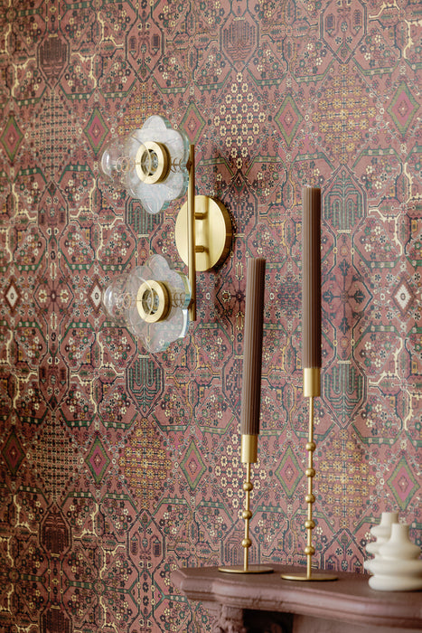 Brass wall sconce with clear glass flower-shaped shades mounted on patterned vintage-style wallpaper, styled above a mantel with decorative brass candlesticks.