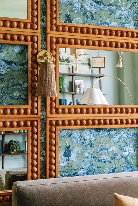 Decorative wall with carved wood-framed mirrors set against patterned blue wallpaper, featuring a brass wall sconce with a tassel shade and reflections of a cozy living space.