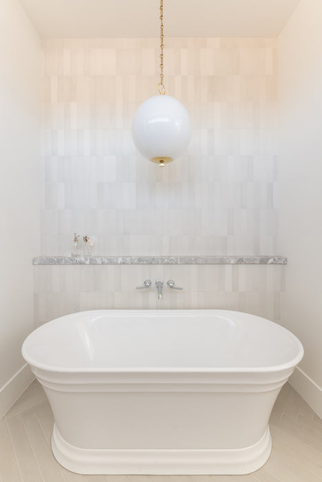 Minimalist bathroom with a freestanding white soaking tub beneath a white globe pendant light with brass chain, set against soft neutral tiled walls.