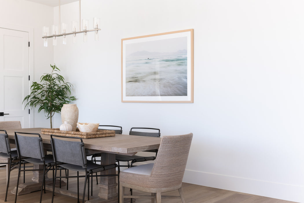 Modern dining room with a wood dining table, mixed seating, minimalist linear chandelier, and coastal artwork on a clean white wall.
