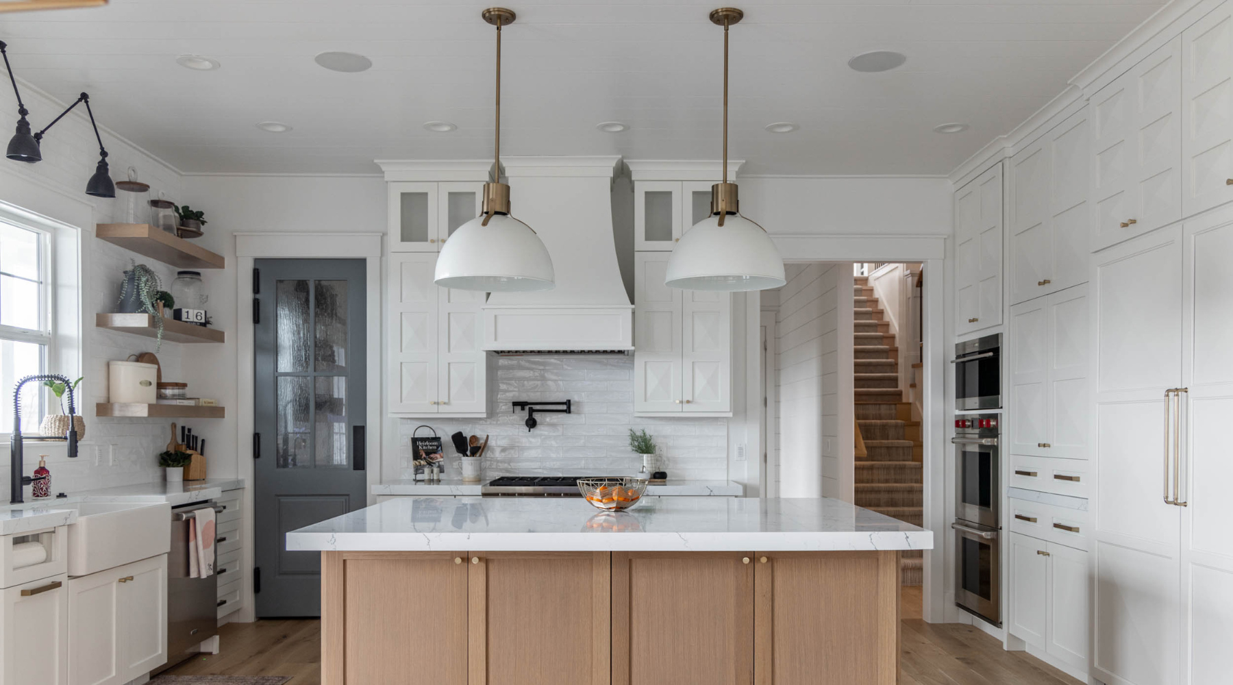 A kitchen with a tan island with white marble countertop, white cupboards, and two handing lights.