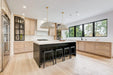 A modern kitchen interior with light wood cabinetry, a black island, white countertops, black bar stools, pendant lights, and large windows with a view of trees.