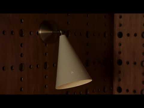 “Wall-mounted sconce with graceful metalwork in a Historic Bronze finish, featuring a Soft Sand-colored shade with a smooth, muted tone. The fixture includes perforated detailing in the metal, adding texture and allowing small points of light to pass through. An adjustable swivel head enables the light to be directed as needed, casting a warm, focused glow while highlighting the sconce’s refined, functional design.
